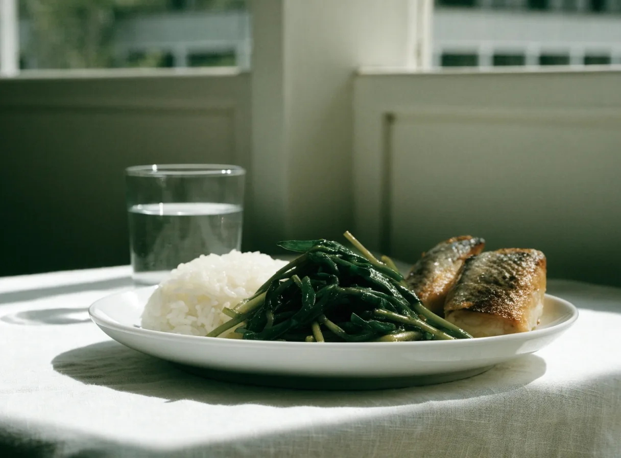 A white plate of rice, stir-fried greens, and grilled fish on linen beside a glass of water